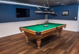 Finished basement remodel in Londonderry NH featuring a custom game room with pool table, TV, wood-look flooring, and modern lighting by Apple Wood Construction.